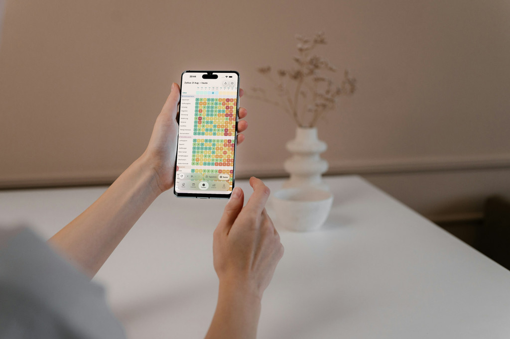 Handen houden een telefoon vast met de kleurgecodeerde symptoomheatmap van PMDD Tracker, boven een witte tafel met een vaas.